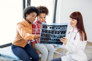 Young female European gp pediatrician, explaining X-ray tomography scan to mixed race girl and her african mom. Mother and daughter looking at CT scan and listening to their family physician
